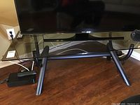 Photo showing the glass top coffee table with black metal frame and angled legs holding a TV, with cables behind the table.