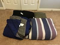 Photo showing folded king size Martel comforter with stripes and blue-gray comforter along with two black and one green throw pillows on carpeted floor.