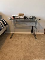 Front view of rectangular metal frame desk with clear glass top against wall, with items on top and beige carpet below.