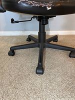 Close-up image of the bottom front portion of the black office chair showing the vinyl tears on the seat and visible adjustment lever and wheels.