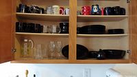 Cabinet interior showing shelves with black dishes, assorted mugs on top shelf, black plates and bowls on the right side, and clear glassware on lower shelves.