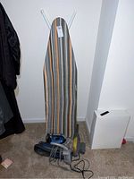 Full lot showing ironing board with striped cover, iron, handheld vacuum, and duster on carpeted floor