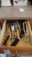 Photo of an open drawer containing assorted kitchen utensils including whisks, spatulas, tongs, peelers, and other small kitchen tools.