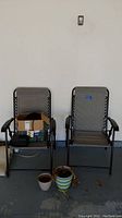 Two gray folding outdoor chairs with box of items on one chair, three plant pots on floor in front