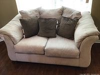 Beige faux leather loveseat with attached back pillows and three dark throw pillows on front seat cushions.