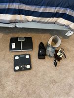 Photo showing two digital scales, one medical foot boot, and one blood pressure cuff on carpeted floor beside a bed.