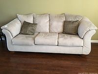Front view of a light beige faux suede sofa with three seat cushions and three back cushions. Two darker taupe decorative throw pillows are on the sofa. Slight wear, some soiling visible on cushions and fabric.