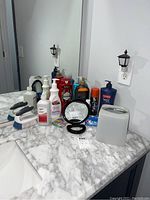 View of countertop with various personal care items including soap, lotions, alcohol, deodorant, toothpaste, tissue box cover, hand mirror, night light, and cleaning brushes.