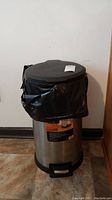 Front view of a stainless steel step trash can with black plastic base and pedal, a black plastic bag liner is visible inside.
