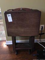 Front view of four wooden TV trays folded and neatly placed in the stand. The tray tops show some wear and aging marks on the wood surface.