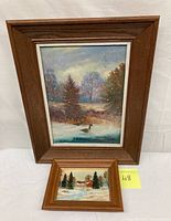 Photo showing two framed winter landscape oil paintings on board, one larger with a duck and one smaller with a rural house and snowy path.