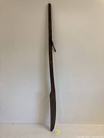Full length view of the antique wooden boat oar showing the blade and shaft against a wall.