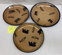 Three Monroe salt glaze pottery plates featuring black bear and salmon silhouettes on tan stoneware with blue rims