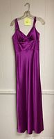 Full length magenta satin evening dress hanging on a clear plastic hanger, wall background.