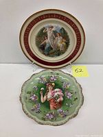 Two decorative portrait plates displayed standing: one round with maroon border and classical woman with children painting, one scalloped green plate with woman holding bouquet and surrounded by flowers.