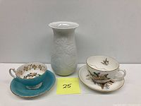 White embossed vase with floral pattern, flanked by two porcelain teacup and saucer sets, one turquoise with gold flowers, the other decorated with flying ducks and gold trim.