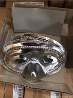 Silver plastic domino mask with ridged decorative edges, laying open in a cardboard box.