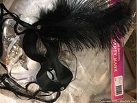 Black party mask with black feathers and attached elastic band, shown with original packaging labeled 'Party Mask'.