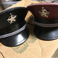 Two military style peaked caps, black and red crowns, with silver star badges and decorative cords on a cardboard surface.