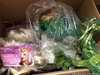 Photo showing various mythological-themed headpieces including a blonde wig with unicorn horn, green Medusa latex piece, and green leafy fairy wreaths packaged together.