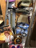 Photo showing packaged Baggins wig ears, Dumbledore hats, Eleven brooch, Hermione wands, and various glasses in original packaging; items stacked on shelving.