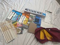 Wide shot displaying all art supplies including watercolor and acrylic paint sets, colored pencils, paint brushes, sketchpad, pencil bag, and message sign.