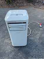 Full front view of white Frigidaire portable air conditioner unit with power cord lying on pavement.