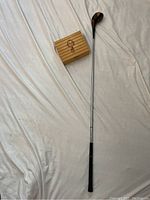 Photo showing vintage wooden-headed golf club and a small wooden box with a golfer engraving on top laid on white fabric