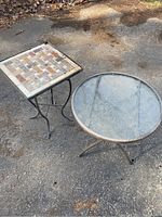 Two patio side tables placed outdoors on a driveway, one with a square tile top and one with a round glass top.