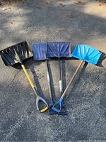 Four snow shovels lying on pavement showing variety of scoop colors and handle types, visible dirt and wear.
