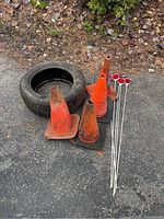 Four orange traffic cones with bases, four roadway reflectors on stakes, and one spare tire outdoors on pavement.