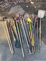 Top-down image showing the entire set of assorted yard tools laid on pavement including pitch forks, shovels, hoes, and trimmer with signs of rust and wear.