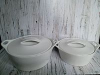 Two white heavy stoneware casserole dishes with lids, displayed side by side against a rustic wood background. Both dishes have curved handles on sides and lids with matching handles.