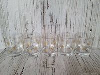 Five clear lowball glasses featuring gold snowflake decorations lined up side by side on a whitewashed wooden surface.