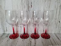 Four red stemmed wine glasses with clear bowls arranged in a row, photographed against a white wood background. Shows the front view of the glasses.