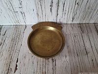 Full view of vintage brass pocket change dish showing round shape and patina on surface, with white rustic wood background.