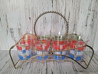 Eight colorful vintage juice glasses arranged in metal wire carrier with coiled handle placed against rustic white wooden background.