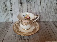 Angled view of Paragon teacup sitting on matching saucer showing ornate goldwork and colorful floral design inside cup and on saucer center.