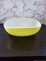 Square vintage PYREX bowl with visible pale yellow exterior and glossy white milk glass interior, placed on dark surface against marble-pattern backdrop.
