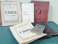 Five German sheet music books and a brown leather satchel laid out flat on a table, showing front covers and condition wear.