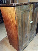 Side angle view of antique wooden cupboard showing wood grain and two paneled doors with ceramic knob.