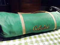 Green cylindrical canvas carry bag with dual straps and yellow script "Cabela's" logo.