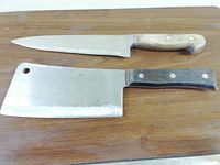 Full view of two cutting utensils laid on wood surface: a slicing chef's knife with wooden handle and a large cleaver with darker handle.