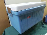 Blue Coleman cooler with white lid, metal clasps on sides and metal handles, showing wear and use marks.