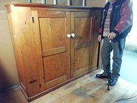 Front left angled view of the pine cupboard showing two doors and white ceramic knobs alongside person for scale.