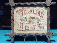Full view of the framed needlepoint sampler showing the complete "Welcome Home" text and house design within a rustic wooden frame.
