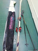 Photo showing the full length of the Kneissl skis with bindings, two black and silver ski poles, and black Salomon ski tote bag with white top section.
