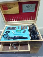 Wide shot of the wooden box opened showing the electric tool, drill bits stored in lid slots, Kodak lens cleaning paper, batteries, and various accessories neatly arranged in a foam insert with two small compartments below