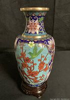 Full view of one cloisonné vase showing bird and floral design on blue background with gold metal rim, mounted on brown wooden stand.