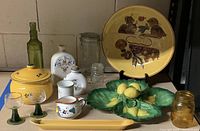 Overview of all collected pottery and glassware showing variety of items and colors including yellow ceramic pot, green leaf dishes with lemons, decorative plates, and glass jars